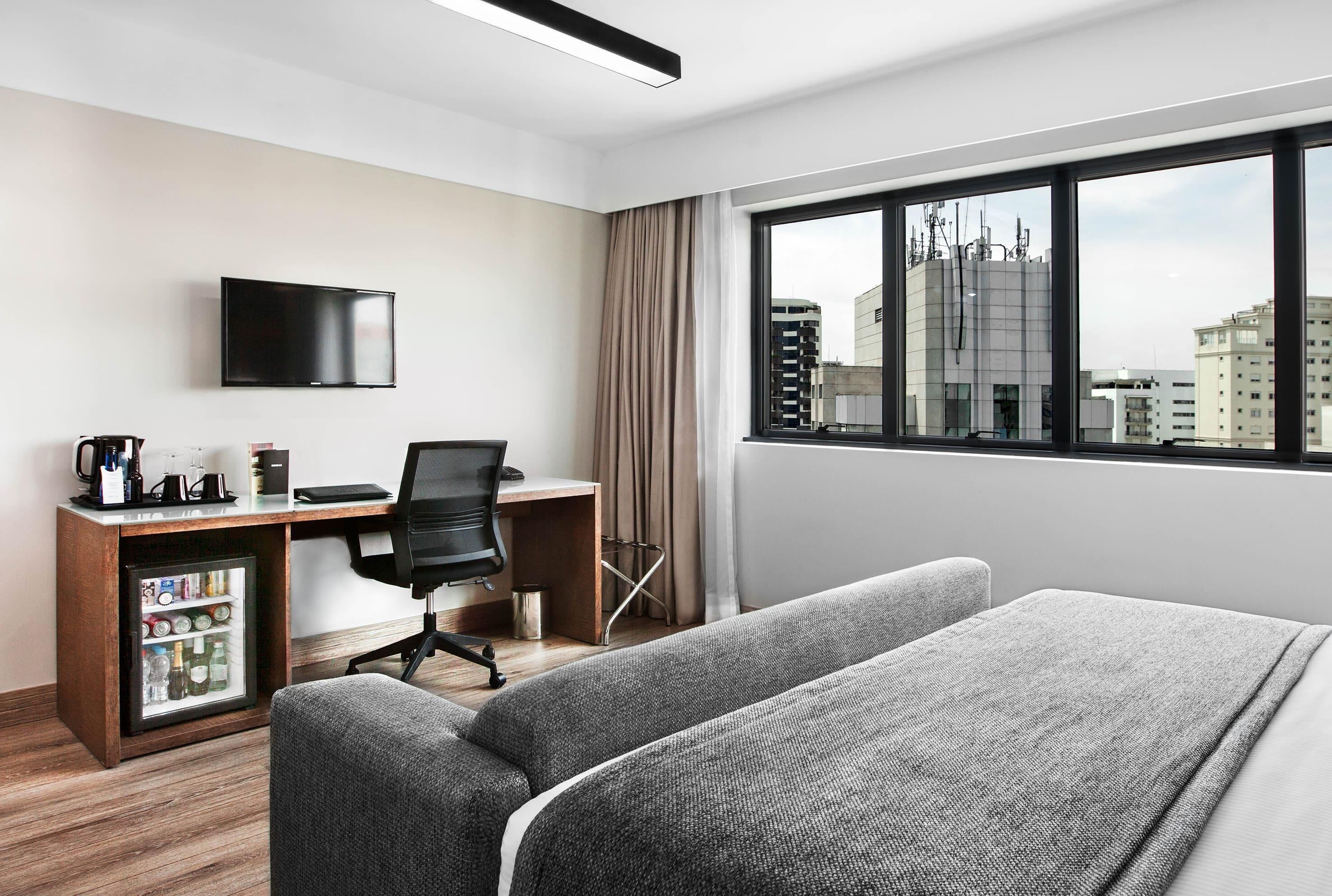 Una habitación de hotel con cama, escritorio con silla, televisor, minibar y ventana con vista a la ciudad.