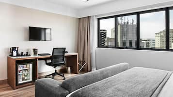 Una habitación de hotel con cama, escritorio con silla, televisor, minibar y ventana con vista a la ciudad.