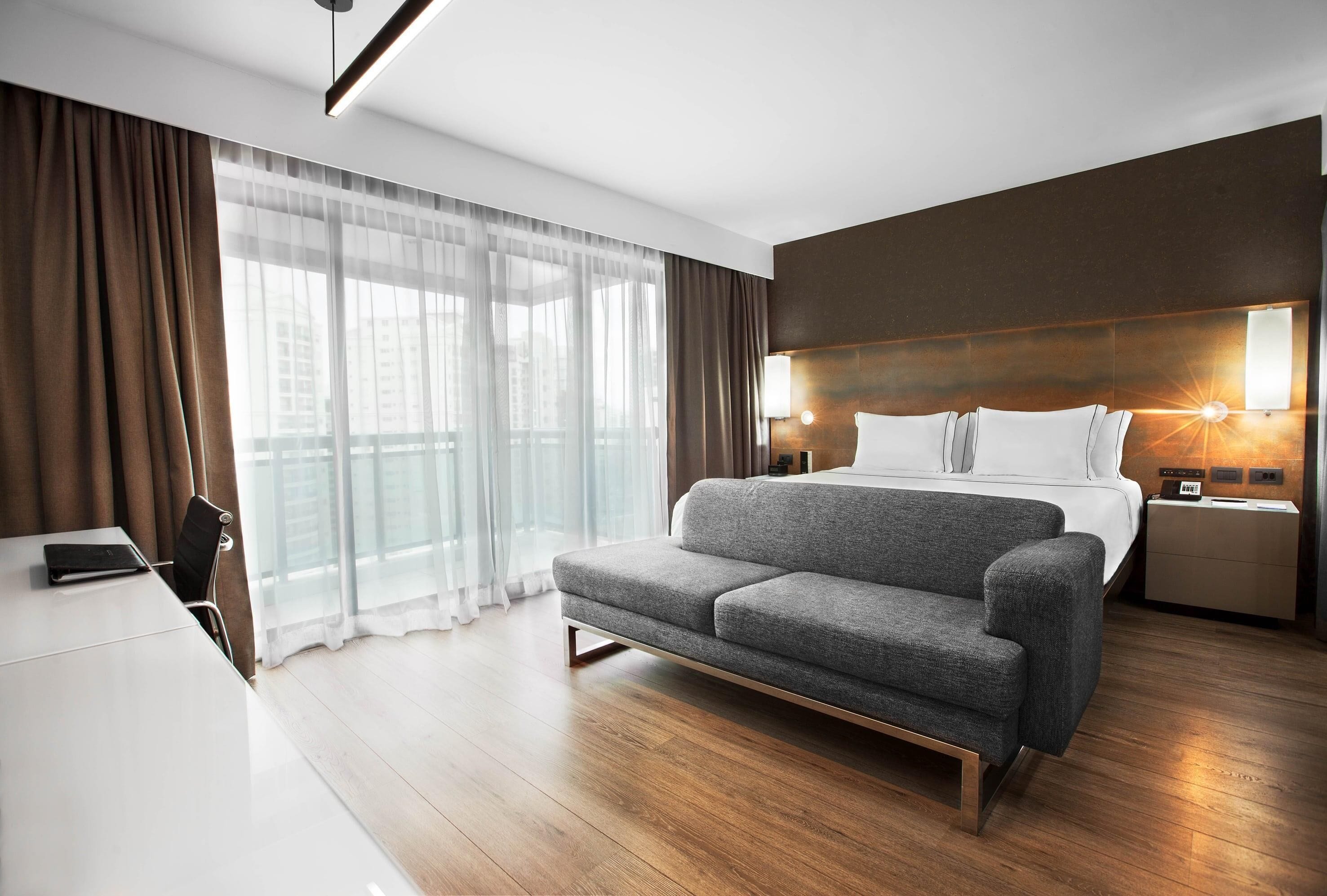 Quarto de hotel moderno com cama-sofá, escrivaninha e uma janela grande com cortinas.