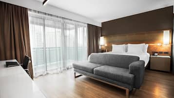 Quarto de hotel moderno com cama-sofá, escrivaninha e uma janela grande com cortinas.