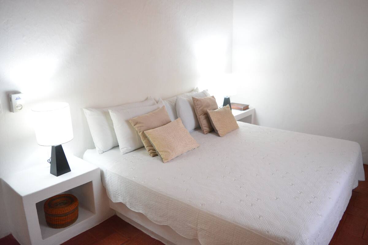 Un dormitorio ordenado con una cama grande, paredes blancas y una mesita de noche con una lámpara y una cesta.