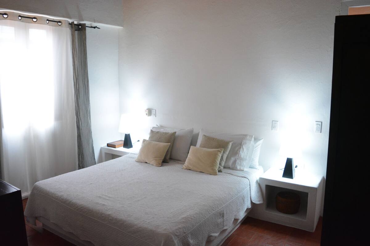 Un dormitorio con una cama grande, dos mesitas de noche, una mesilla auxiliar, una ventana con cortinas y una puerta.