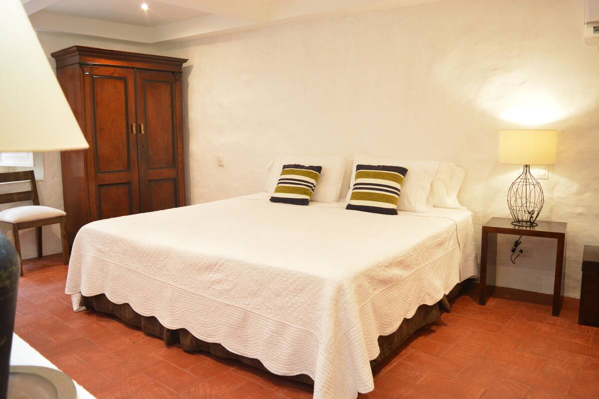 Un dormitorio con una cama grande, un armario de madera, una mesita de noche con lámpara y una silla.