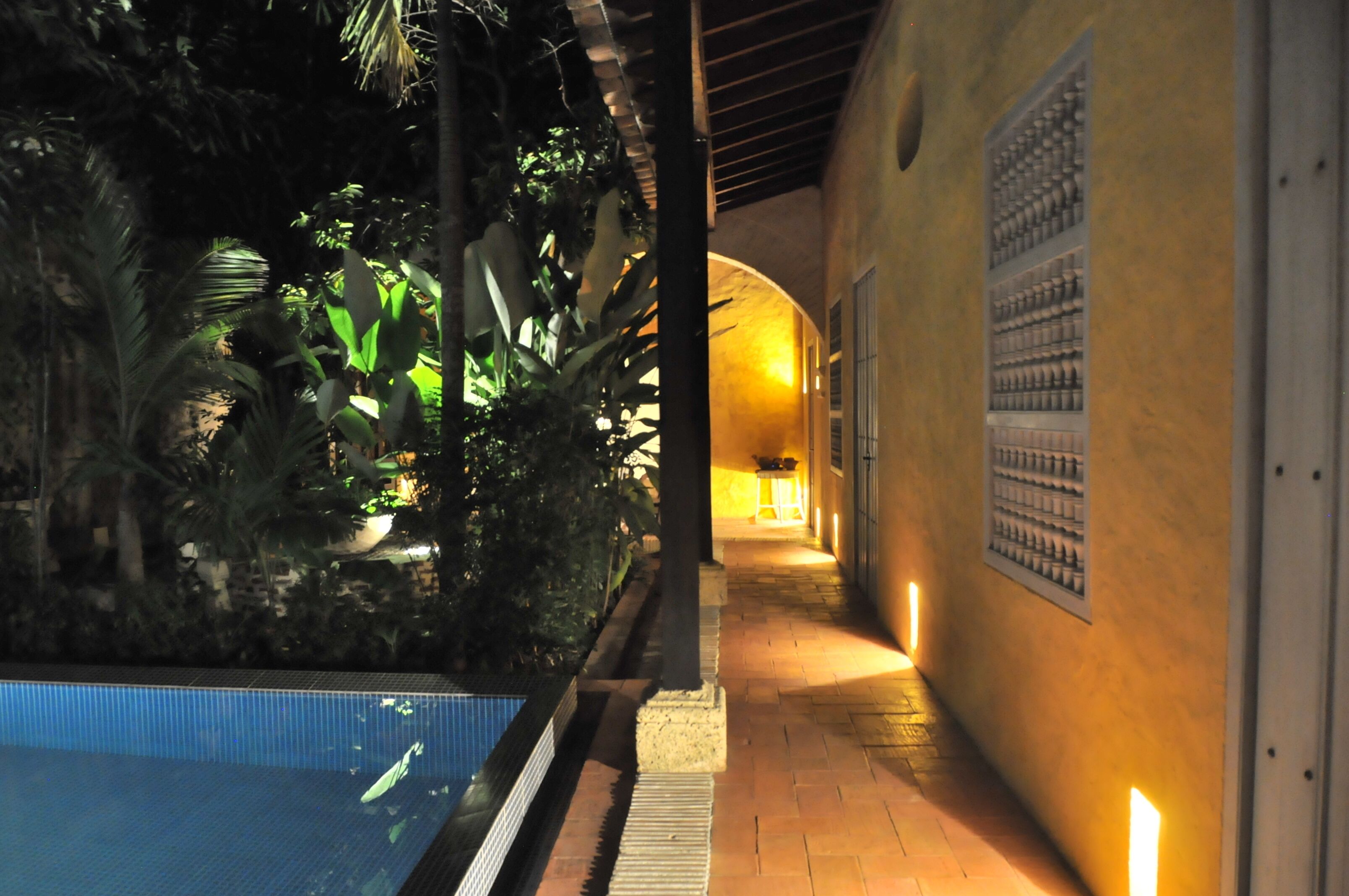 Uma área à beira da piscina com um caminho ladrilhado, palmeiras e um edifício com fachada amarela.