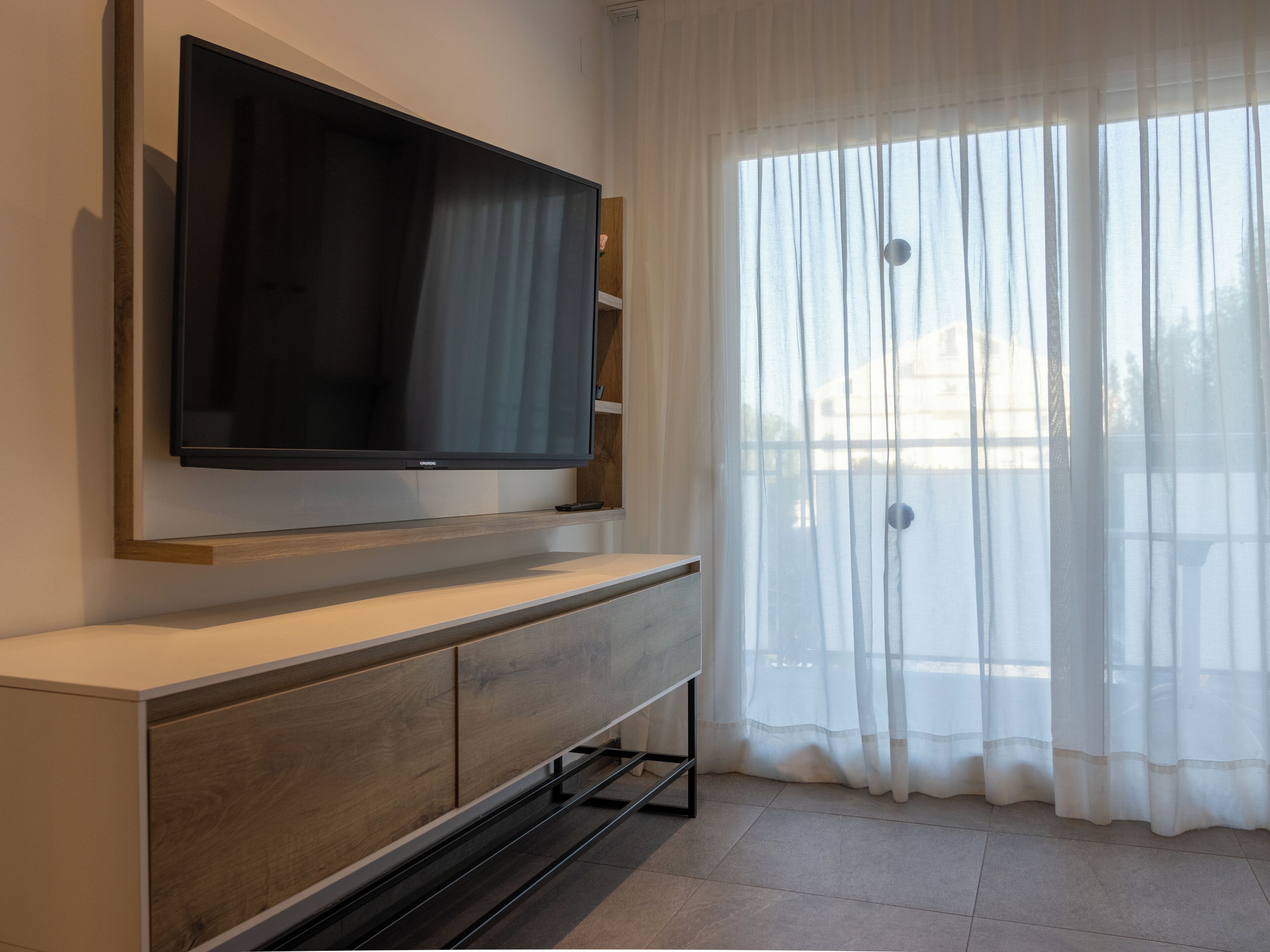 A modern living room with a flat-screen TV mounted on a wooden console, a large window with sheer curtains, and a tiled floor.