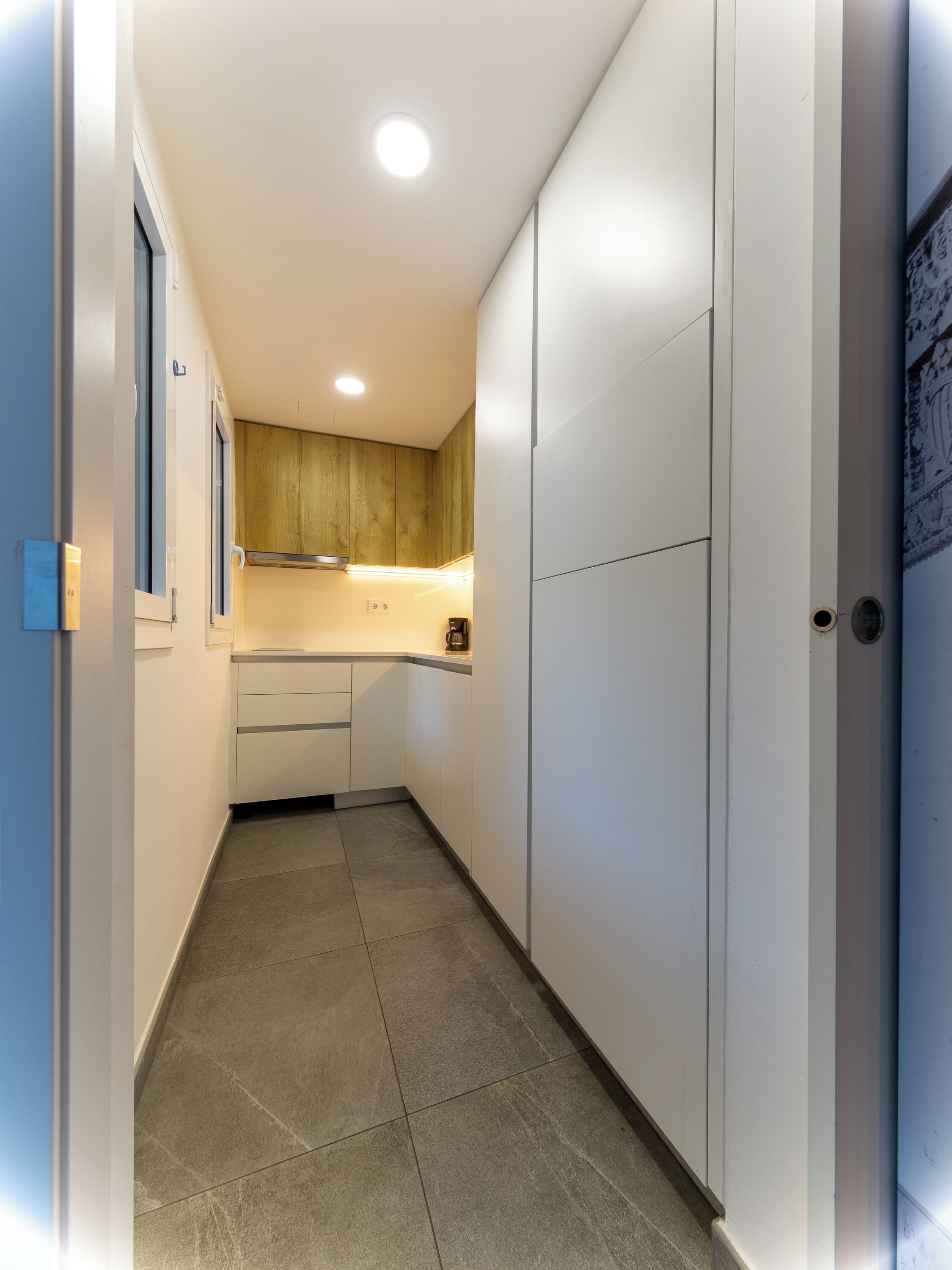 Un couloir étroit donnant sur un espace cuisine avec des meubles blancs, un plan de travail en bois et un sol carrelé.