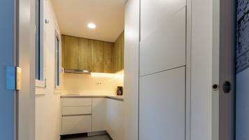 Un couloir étroit donnant sur un espace cuisine avec des meubles blancs, un plan de travail en bois et un sol carrelé.