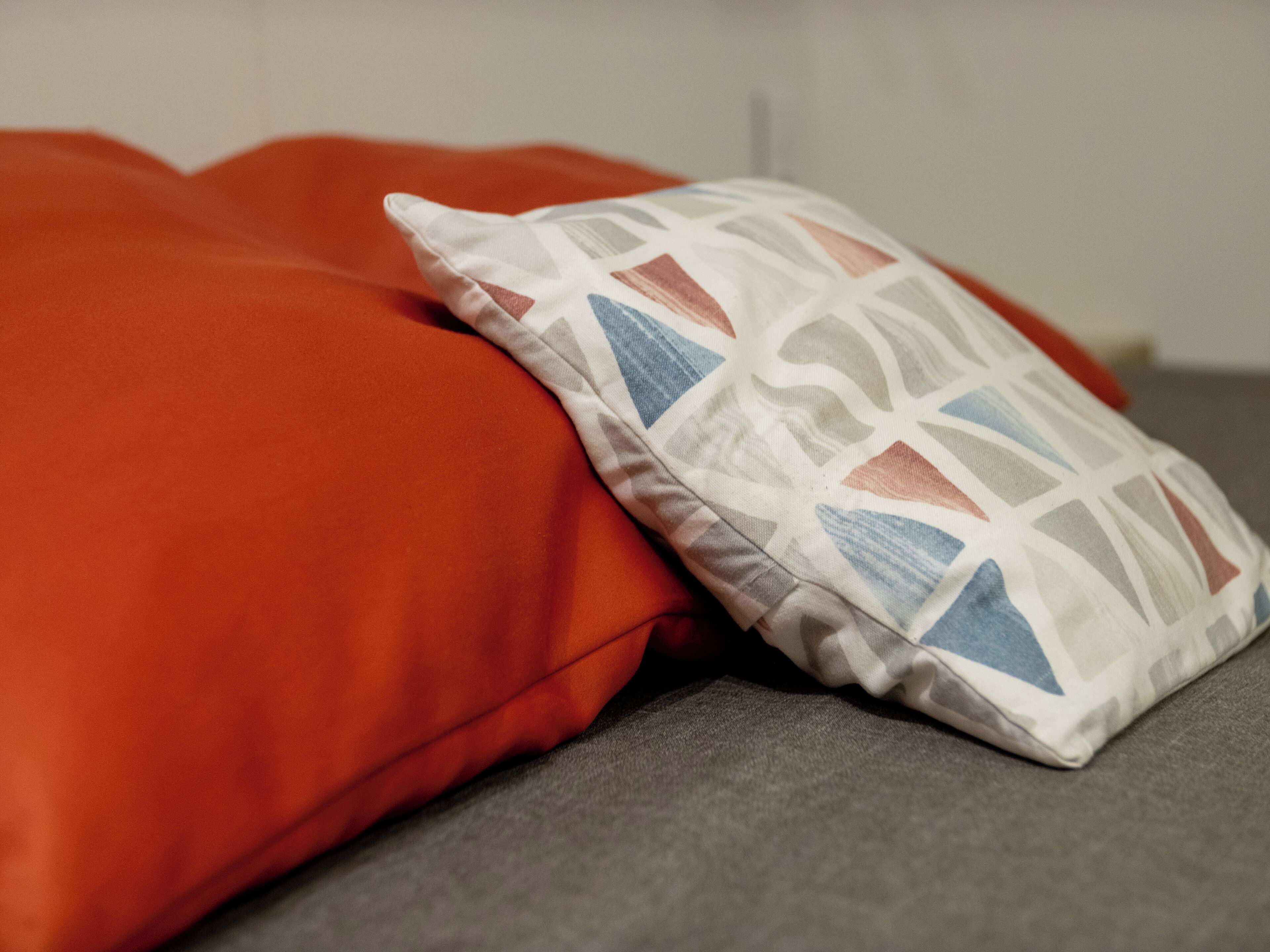 A bed with a grey mattress and two pillows, one orange and one with a geometric pattern.