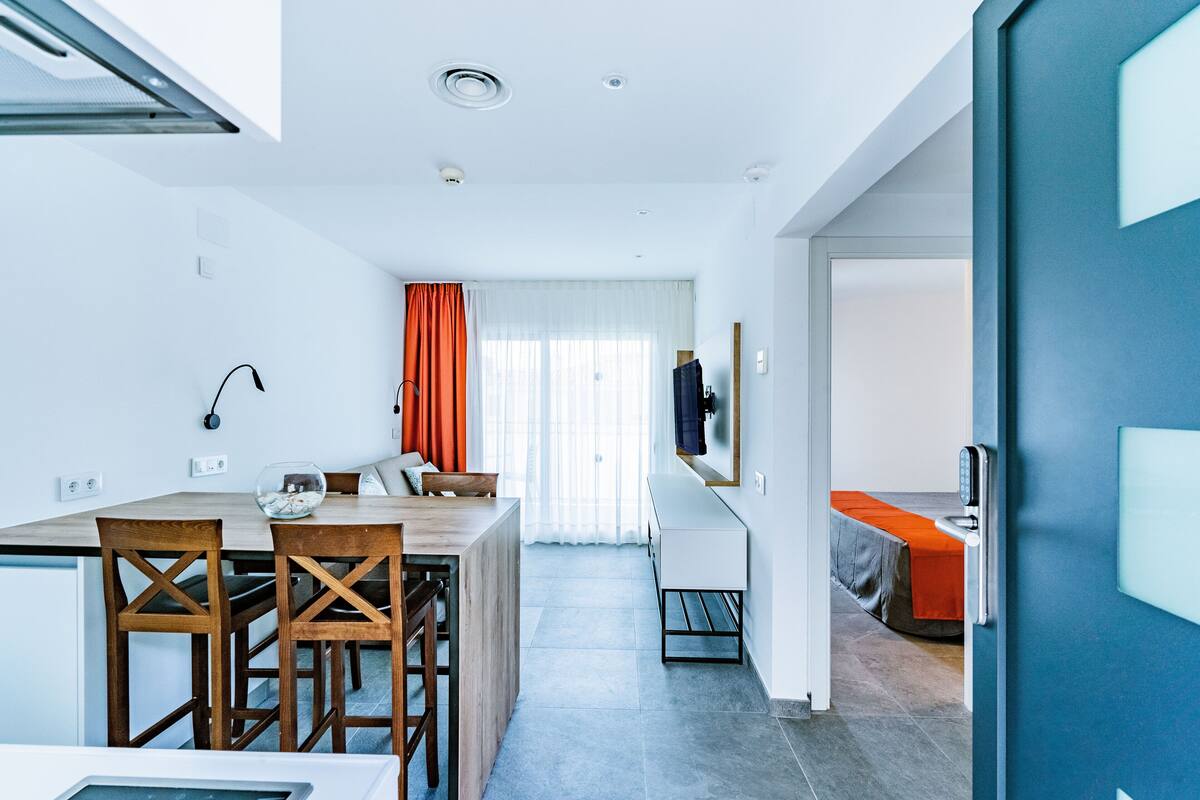 Una habitación de hotel moderna con zona de comedor, una cama con colcha naranja y un ventanal con cortinas.
