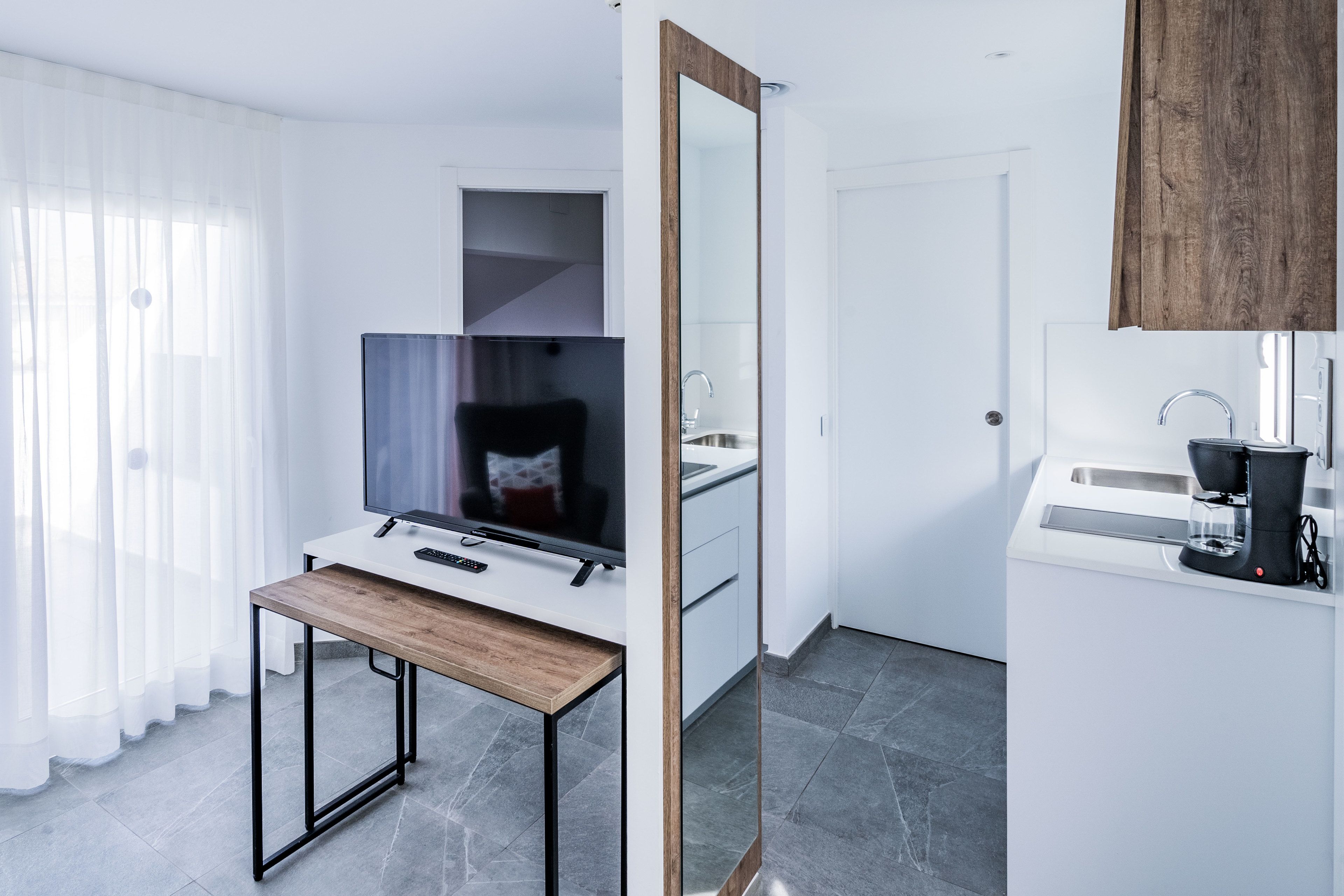 A modern hotel room with a flat-screen TV, a wooden table, a large mirror, a kitchen area with a coffee machine, and a white wardrobe.