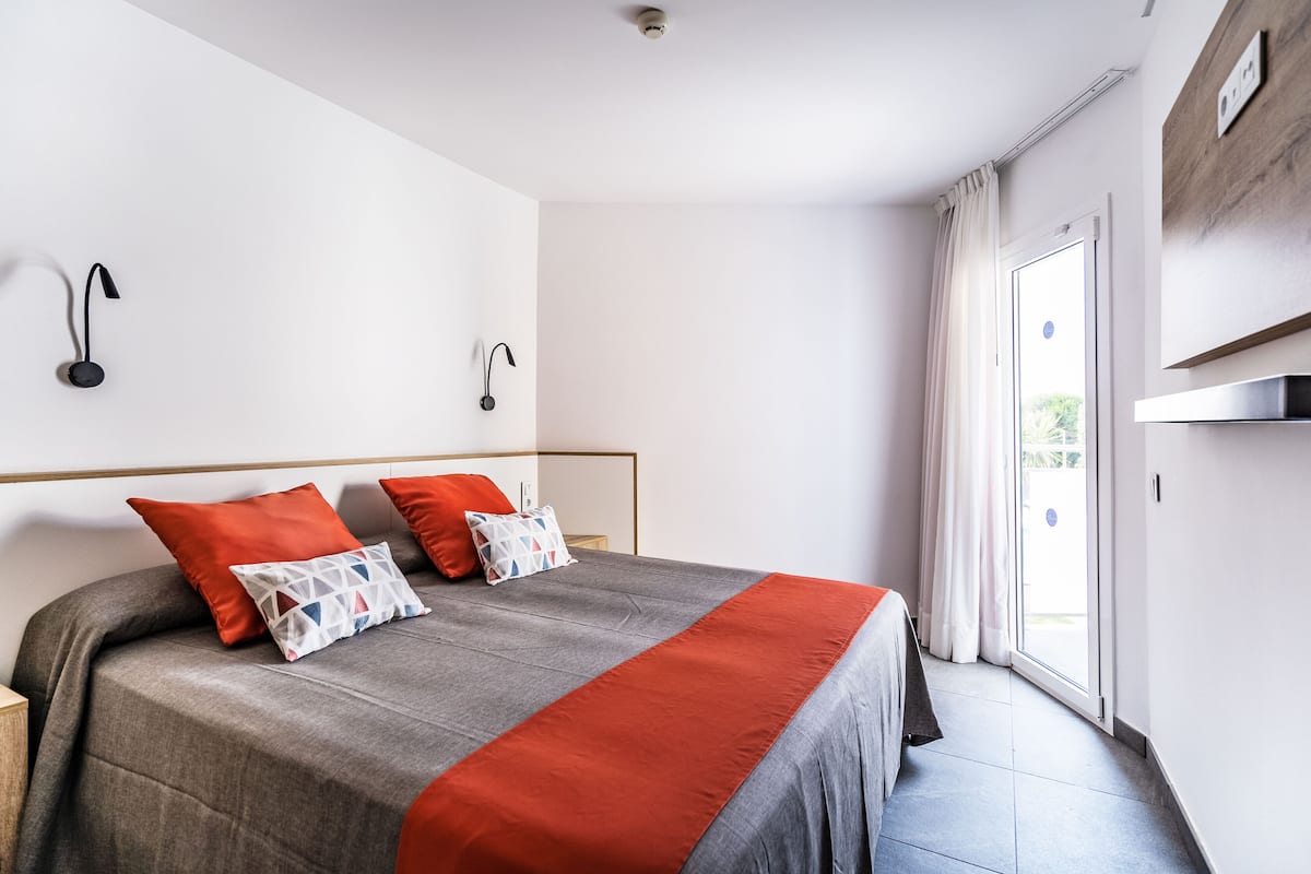 A modern hotel room with a large bed, two wall-mounted reading lamps, a sliding glass door to the balcony, and a wooden panel on the wall.