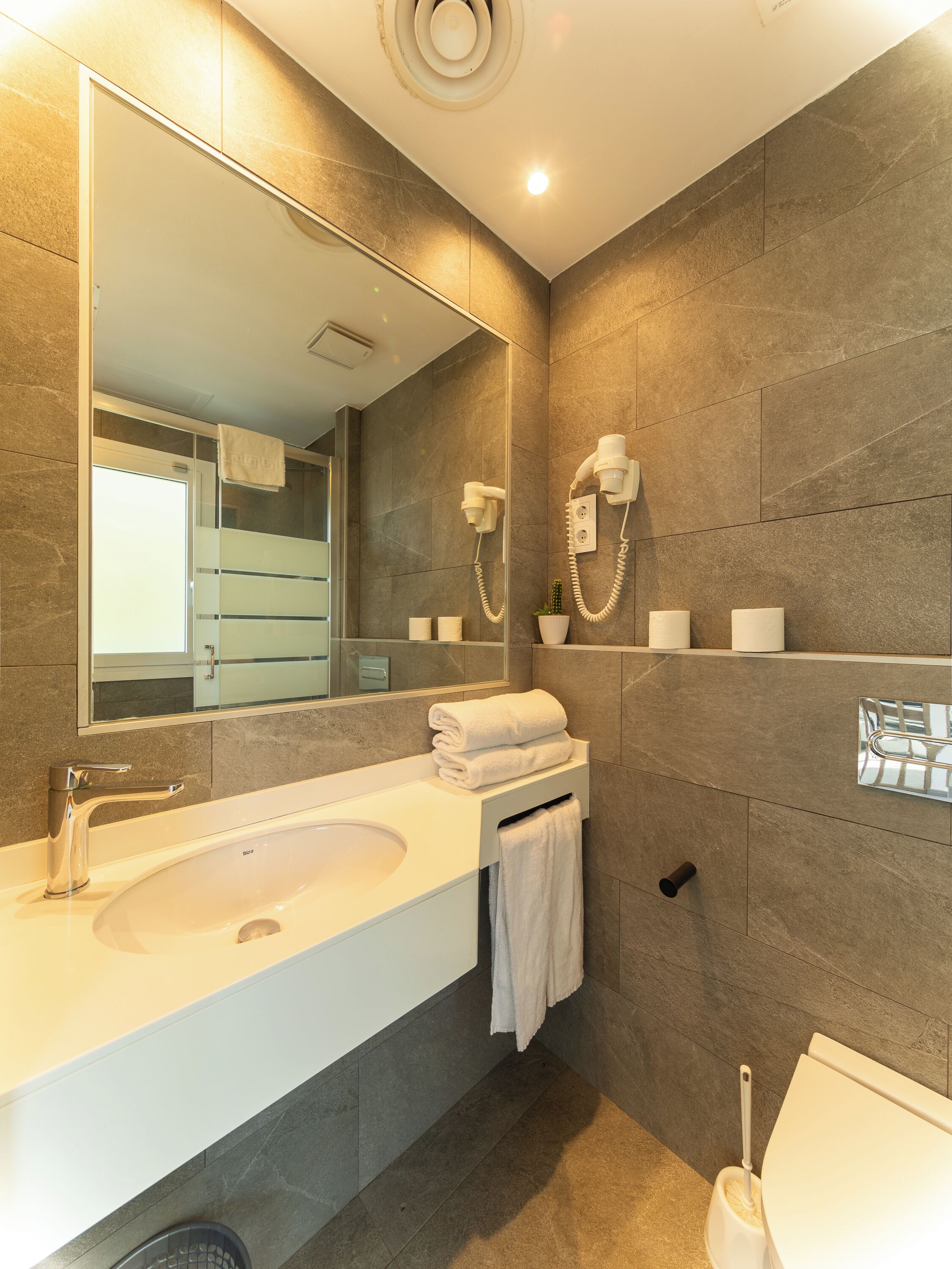 A modern bathroom with a large mirror, a sink, a toilet, and a hairdryer mounted on the wall.