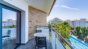 A balcony with a glass door, a brick wall, a round table with chairs, and a view of a swimming pool and buildings in the background.