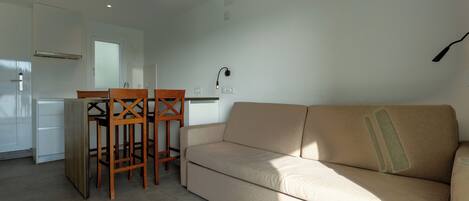 Una sala de estar con un sofá beige, sillas de madera, una mesita y una zona de cocina con refrigerador y armarios.