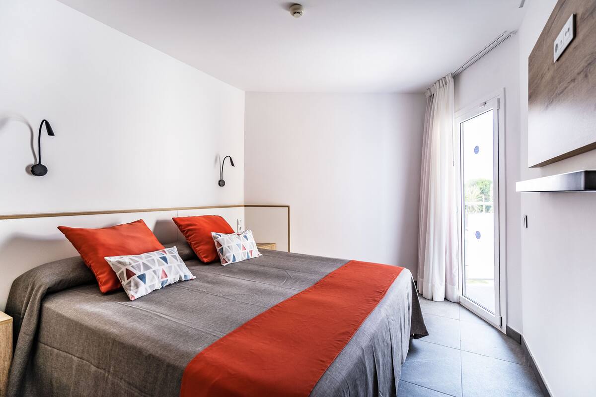 Habitación de hotel moderna con una cama grande, dos almohadas, una manta roja y una puerta corredera de vidrio al exterior.