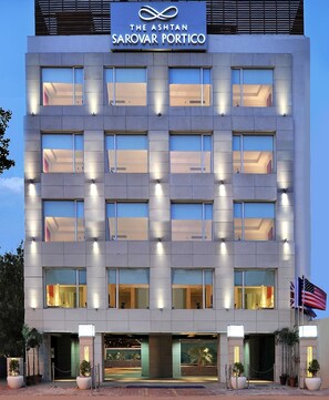 Un moderno edificio de hotel con un letrero que dice "The Ashton Sarovar Portico". Cuenta con varios pisos con ventanas regularmente espaciadas y una entrada bien iluminada.