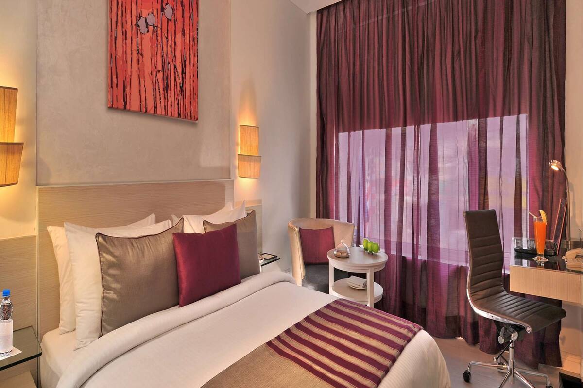 Habitación de hotel con cama, escritorio, silla, una mesita pequeña y un ventanal con cortinas.