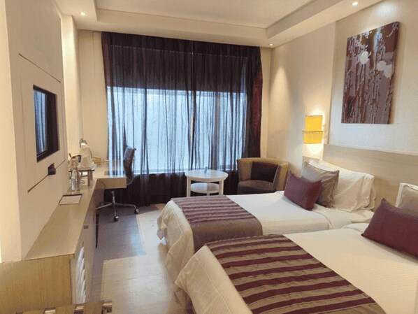 A hotel room with two beds, a desk, a chair, a small table, a TV, and a window with curtains. - The Ashtan Sarovar Portico Delhi (New Delhi)