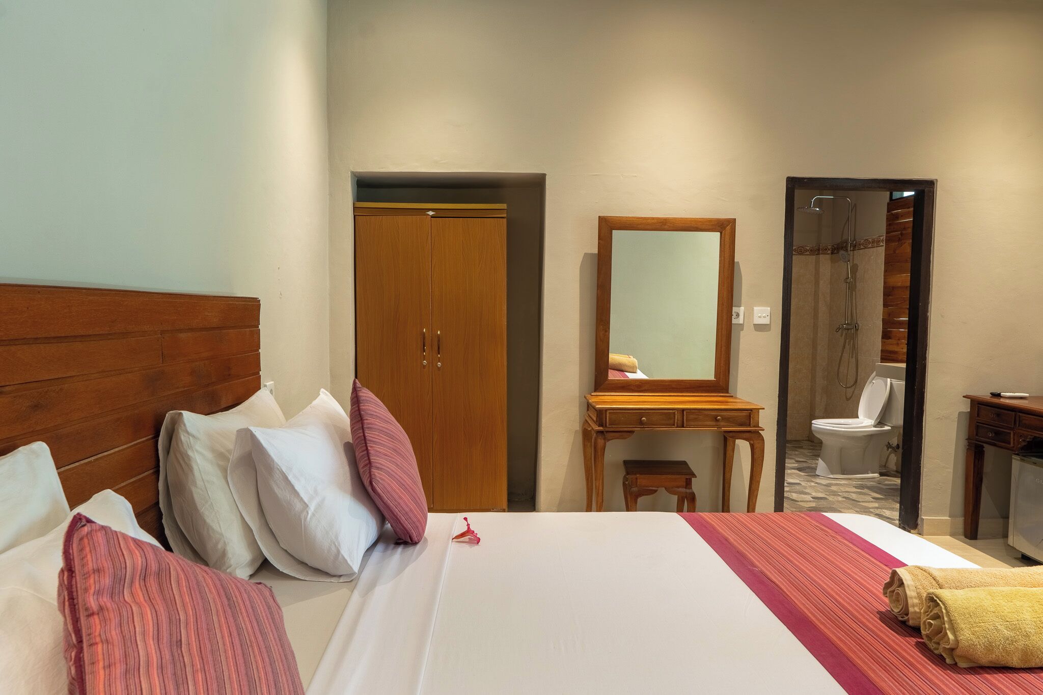 Ein Hotelzimmer mit einem hölzernen Kopfteil, einem Bett mit weißem und rosa Kissen, einer Holzkommode, einem Spiegel und einem durch eine offene Tür einsehbaren Badezimmer.