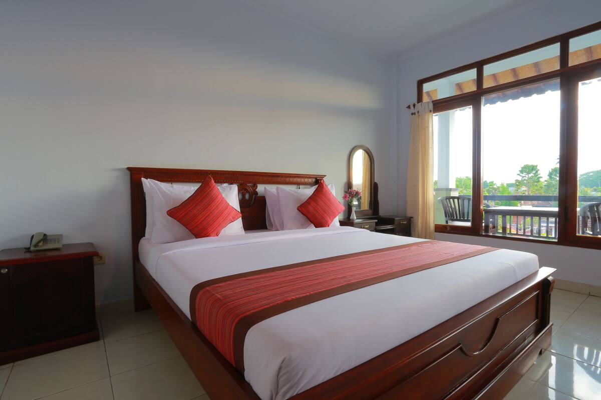 A large bed with a wooden frame and white bedding, a nightstand with a phone, and a window with a view of greenery.