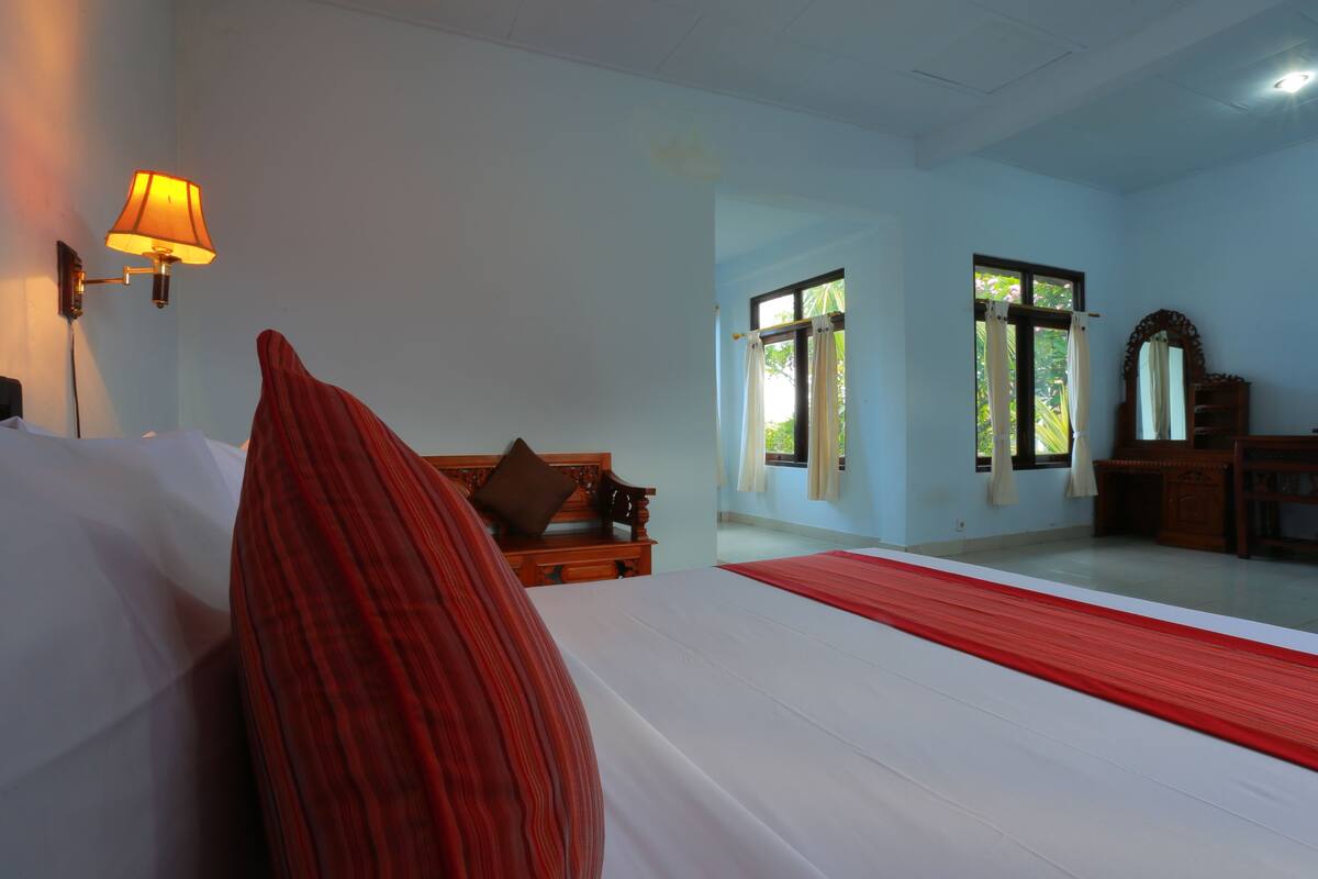 A hotel room with a bed, a wooden bench, a lamp, and a window with curtains.
