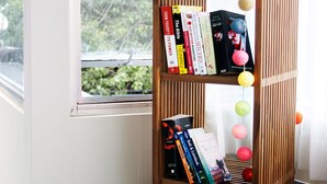 Ein Bücherregal mit Büchern und einem Stoffhasen, eine Tasse und ein Laptop auf einem Tisch neben einem Fenster.