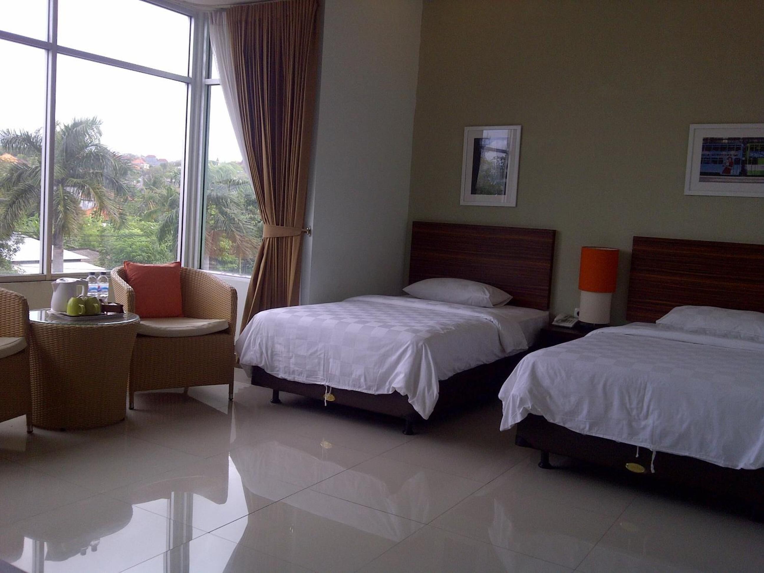 A hotel room with two beds, a sitting area with a chair and table, and large windows with curtains.
