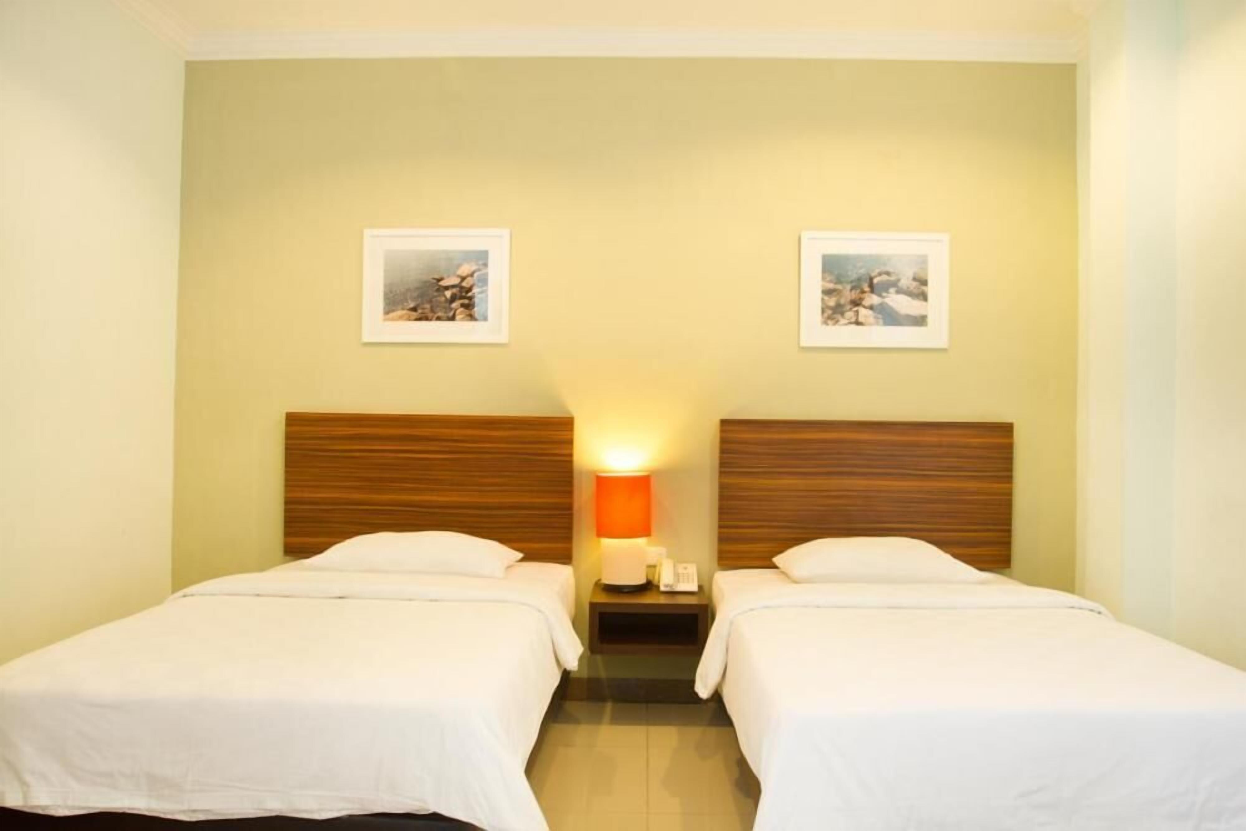 A hotel room with two single beds, a wooden headboard, a nightstand with a lamp, and two framed pictures on the wall.