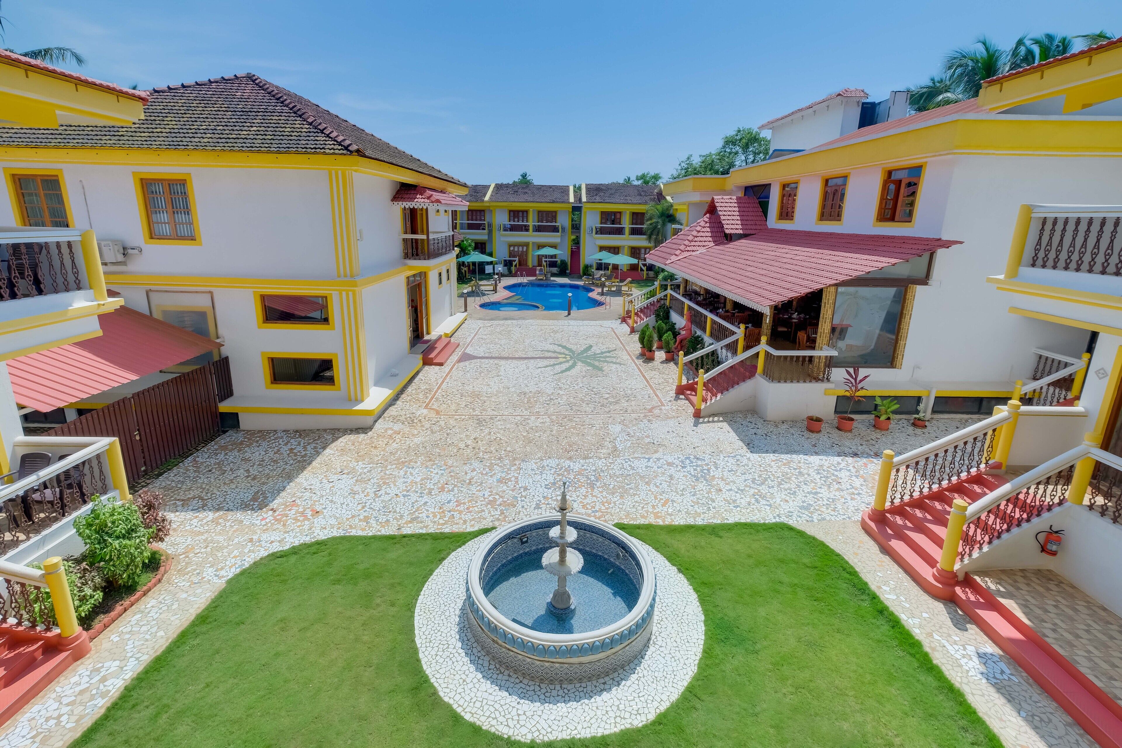 A courtyard with a central fountain, surrounded by multi-story buildings with balconies and a swimming pool in the background.