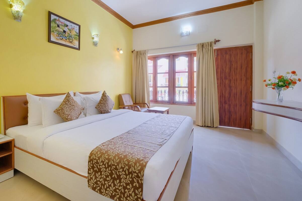 Une chambre d’hôtel avec un grand lit, une chaise, une petite table, une fenêtre avec des rideaux et un luminaire fixé au mur.