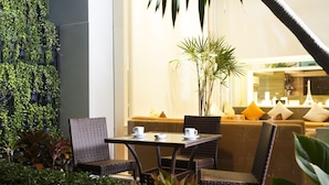 Un área de estar al aire libre con muebles de mimbre y una mesa, rodeada de exuberante follaje y una planta grande en maceta.