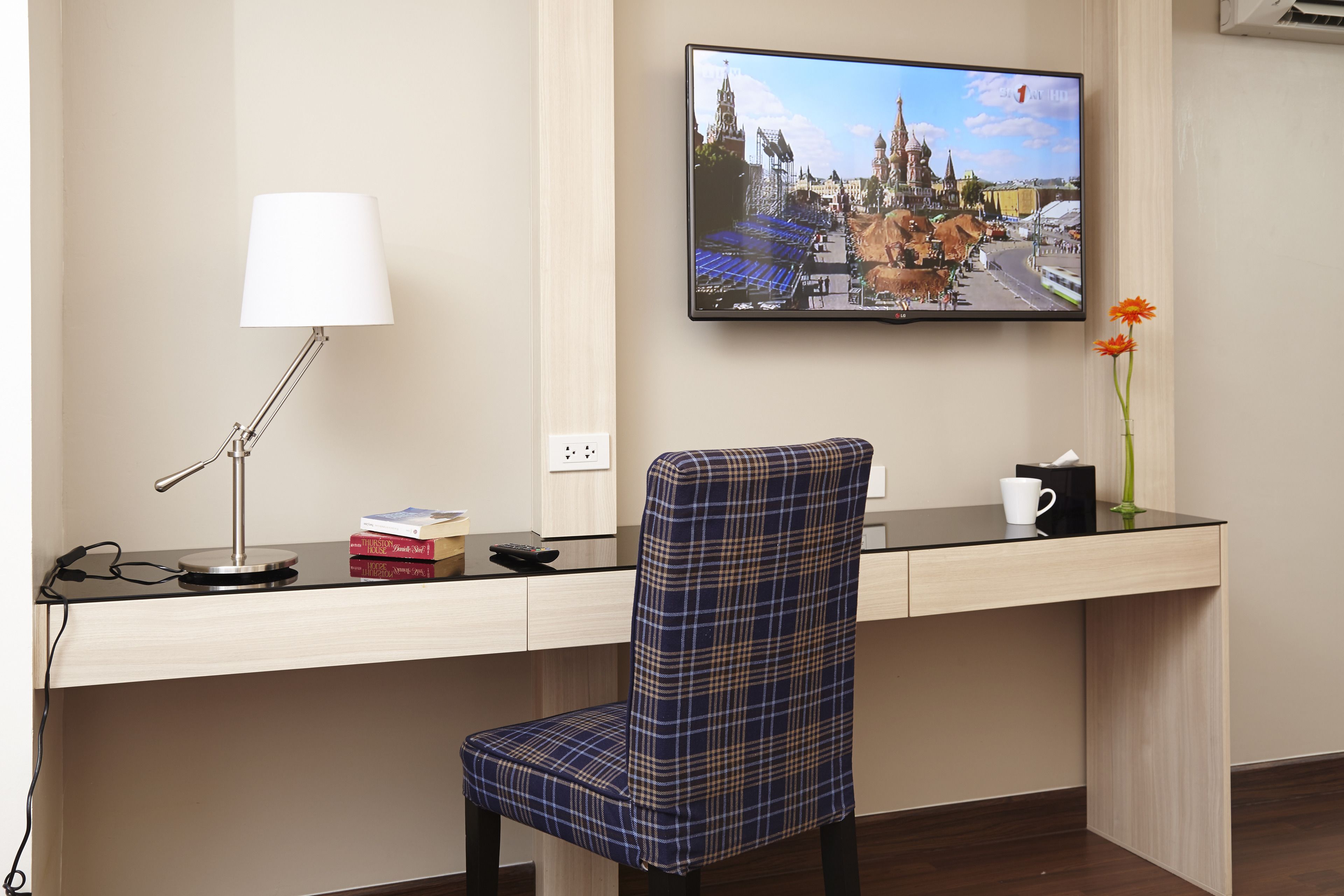 A hotel room with a desk, a chair, a lamp, a flat-screen TV, and a vase with flowers.
