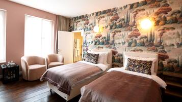 Quarto de hotel com duas camas, área de estar com uma poltrona, uma pequena mesa com garrafas e um mural na parede.