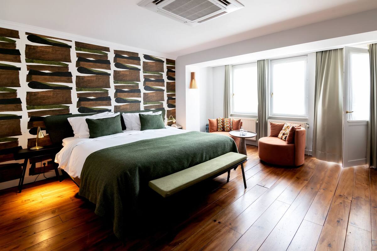 A modern bedroom with a wooden floor, a large bed with a green bedspread, a bench, a chair, and a wall with a unique wooden panel design.