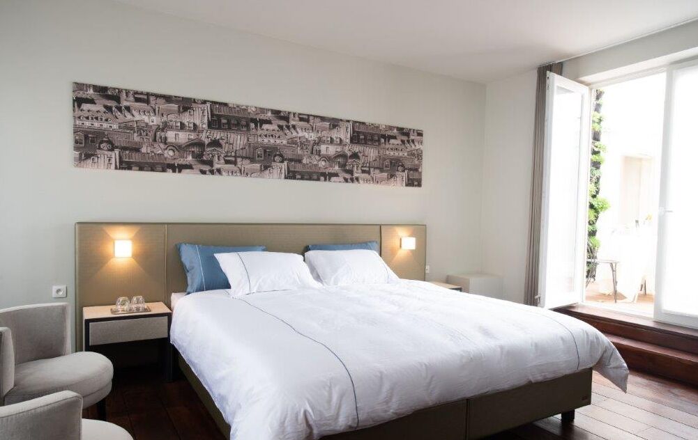 A hotel room with a large bed, a headboard with wall-mounted lights, a side table with a glass, and a view of the outdoors through a large window.