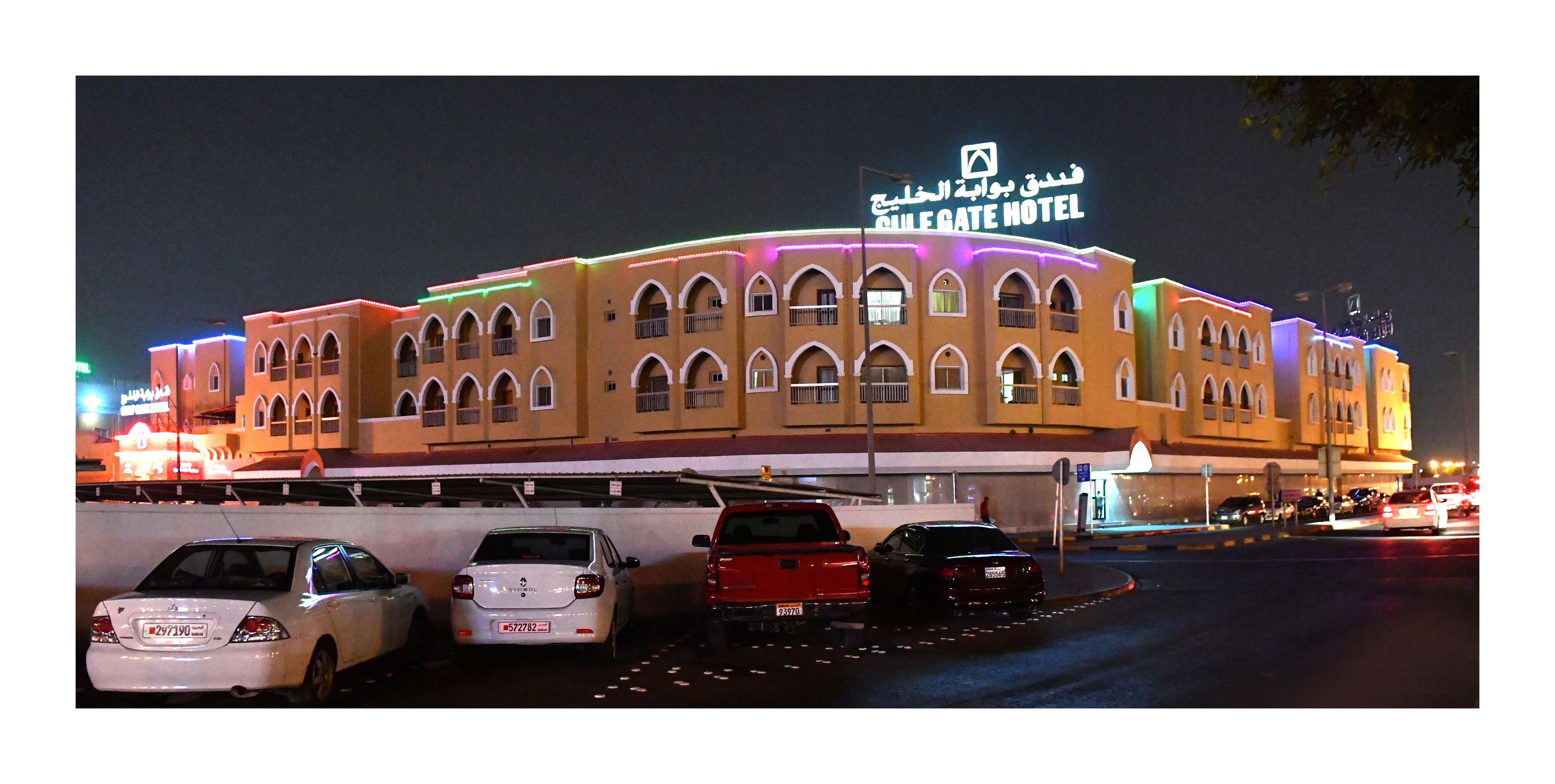 Een hotel met een bord in Arabisch, meerdere balkons, en een parkeerplaats met auto's.