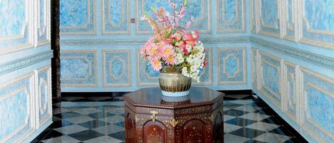 Una habitación con piso a cuadros, una mesita de madera con un jarrón de flores y paredes con diseño azulado.