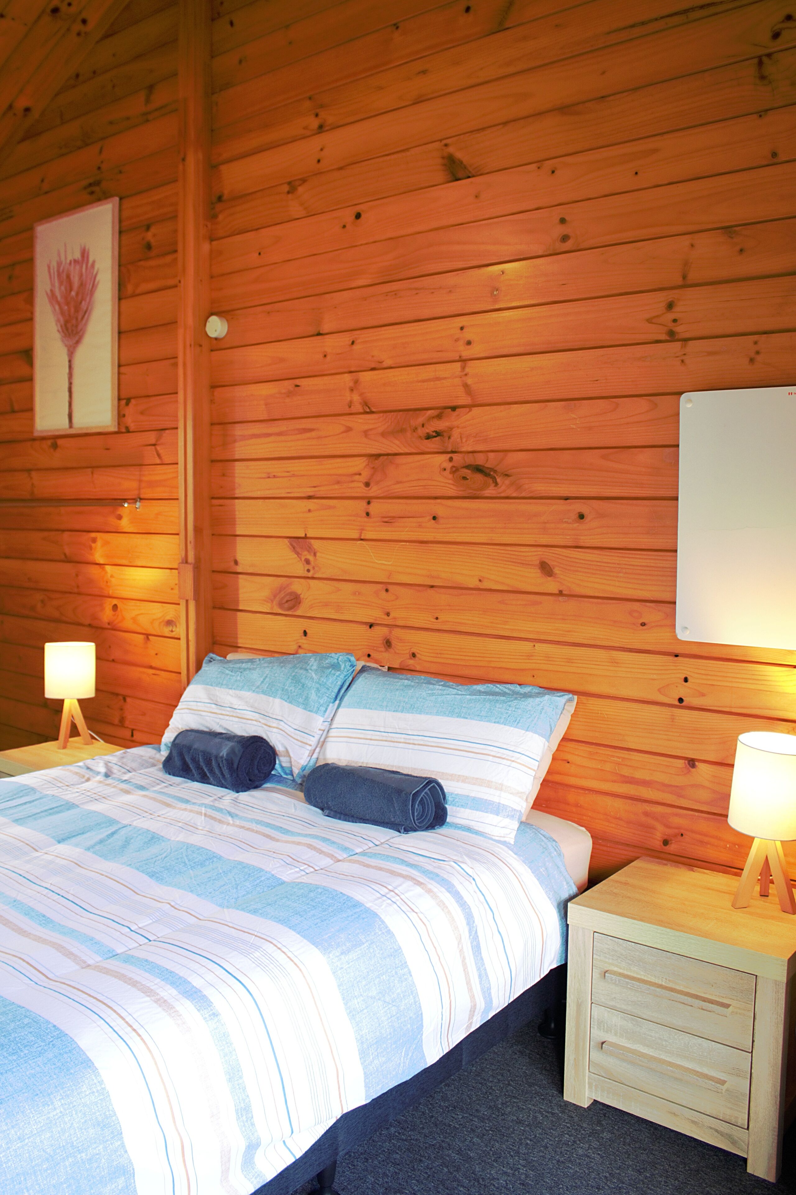A wooden cabin bedroom with a bed, two bedside tables, and a wall-mounted lamp.
