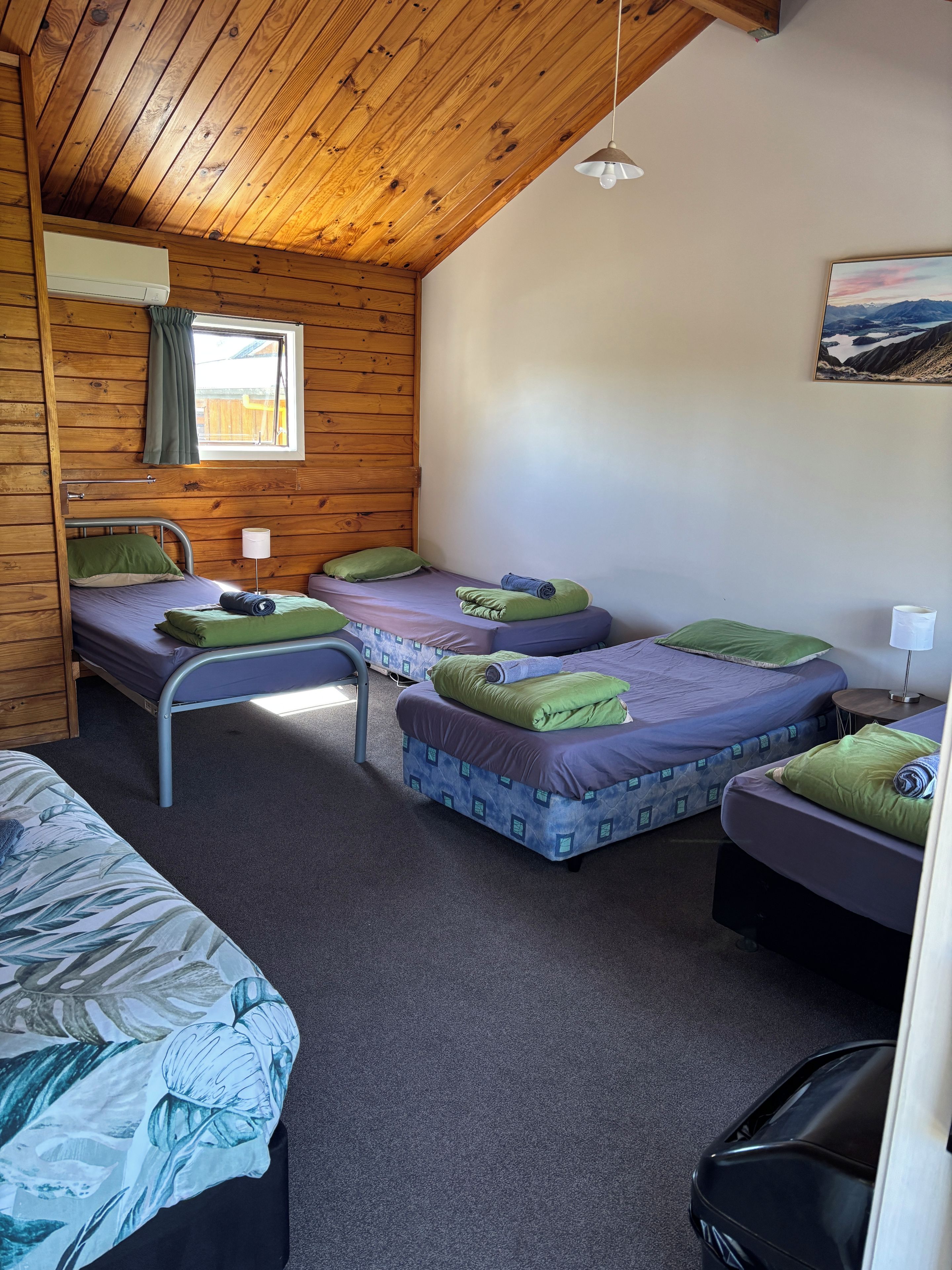 Quarto com paredes de madeira e teto inclinado, com três camas com roupa de cama roxa e verde, uma janela com cortina e um quadro na parede.