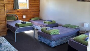 A room with wooden walls and a sloped ceiling, featuring three beds with purple and green bedding, a window with a curtain, and a painting on the wall.