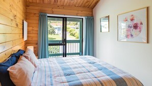 A bedroom with a wooden ceiling, a bed with a plaid bedspread, a window with curtains, and a framed floral artwork on the wall.