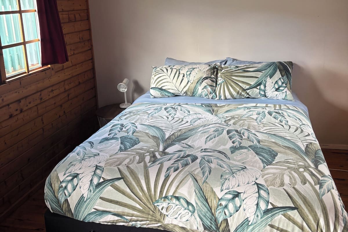 Ein Bett in einem Zimmer mit Holzwänden und einer geneigten Decke, ein Fenster mit Vorhängen und einem Deckenventilator.