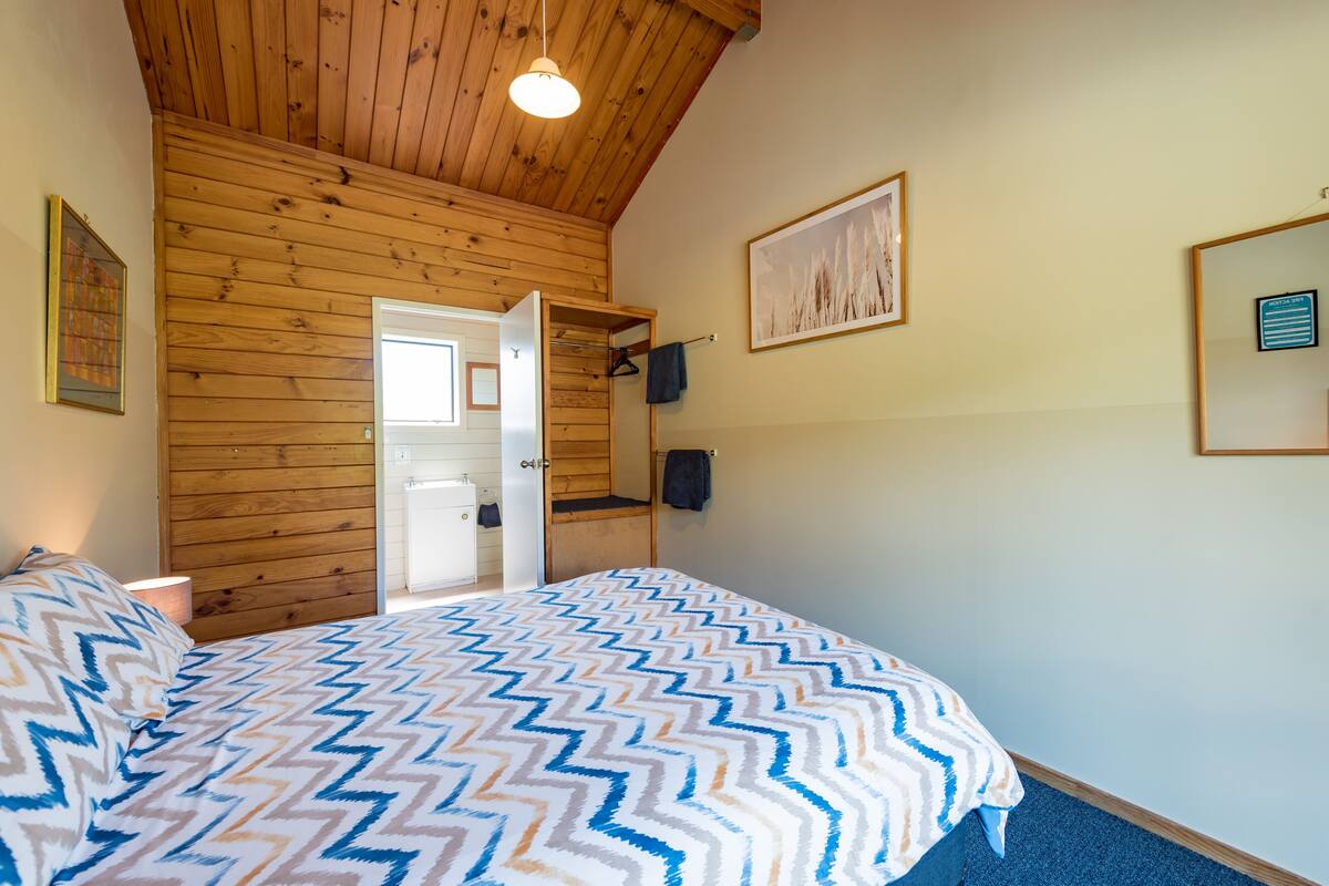 Ein Schlafzimmer mit Holzbalkendecke, einem Bett mit gemustertem Überwurf, einer Tür zu einem Badezimmer und einem gerahmten Bild an der Wand.