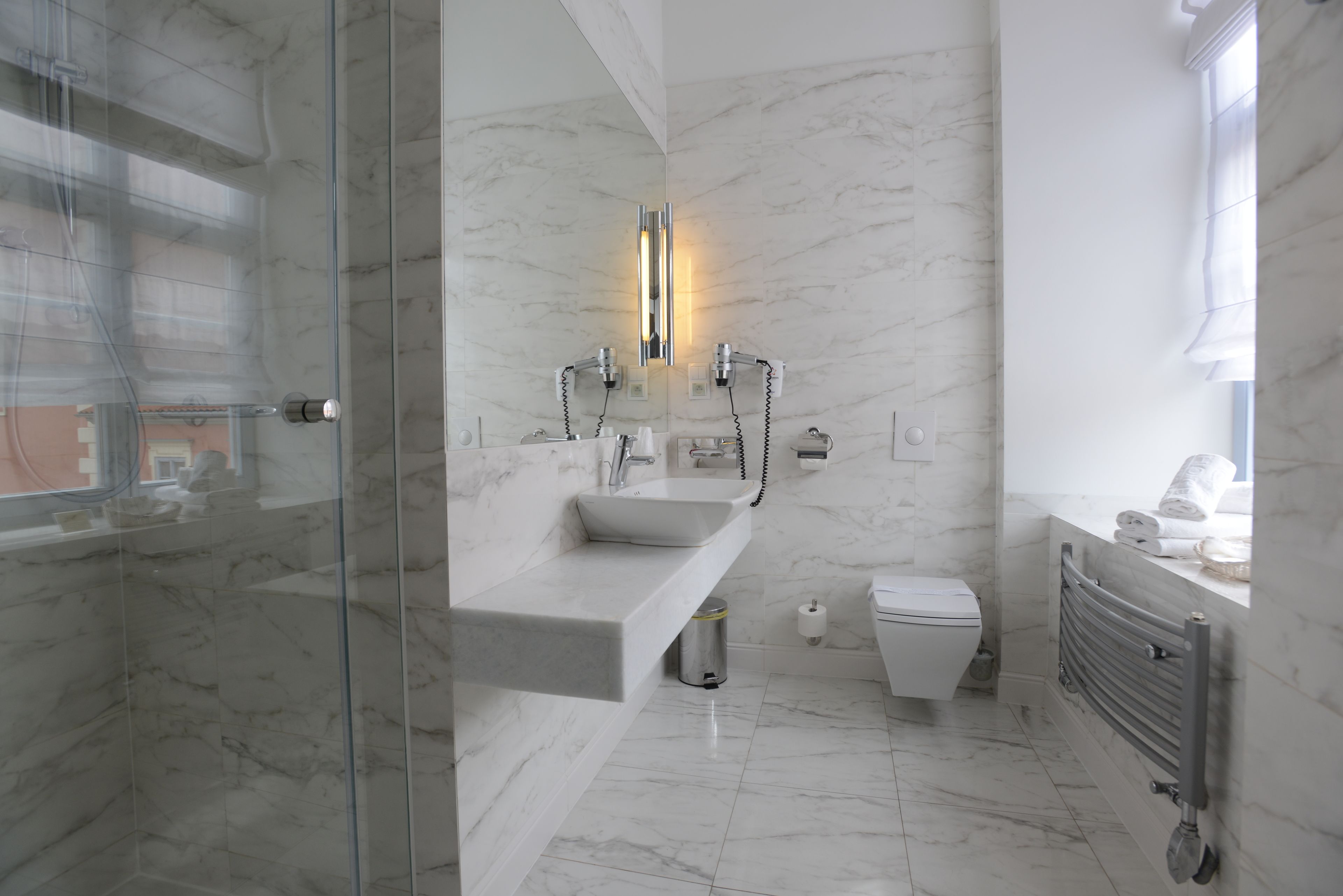 Un bany modern amb plat de dutxa de vidre, terra de rajola de marbre, un lavabo blanc amb taulell de marbre, un vàter i un radiador fixat a la paret.