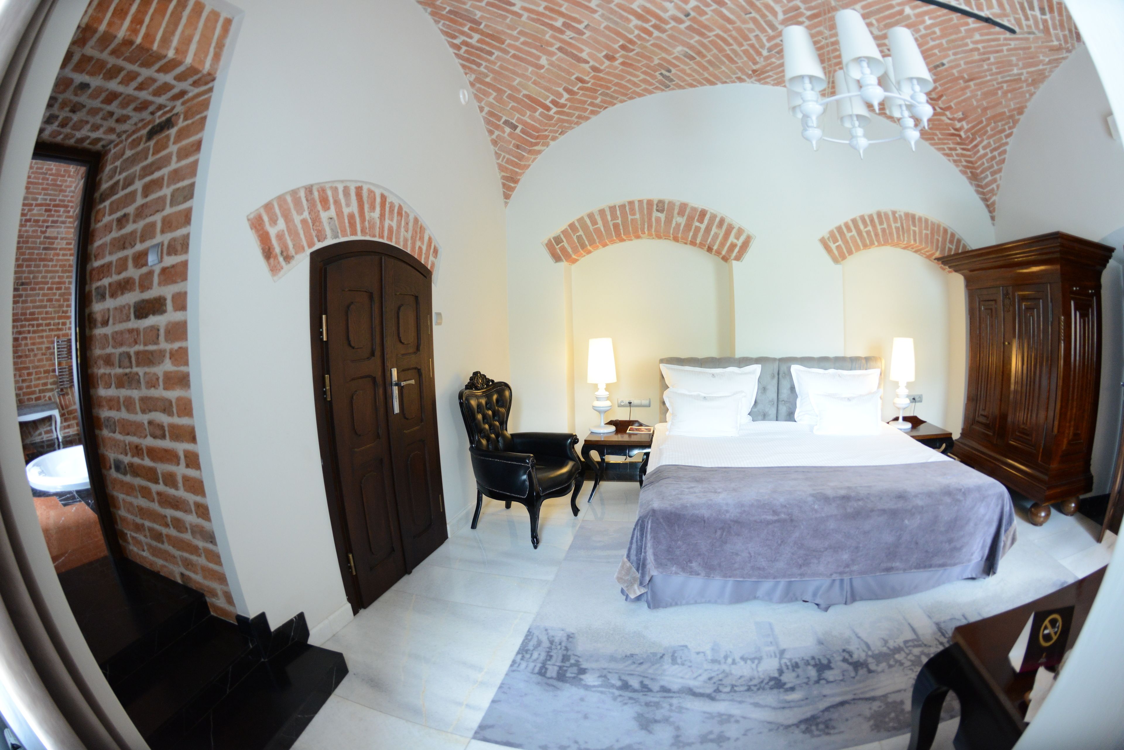A room with a brick ceiling, a bed with a white headboard, a wooden wardrobe, and a small table with a lamp.