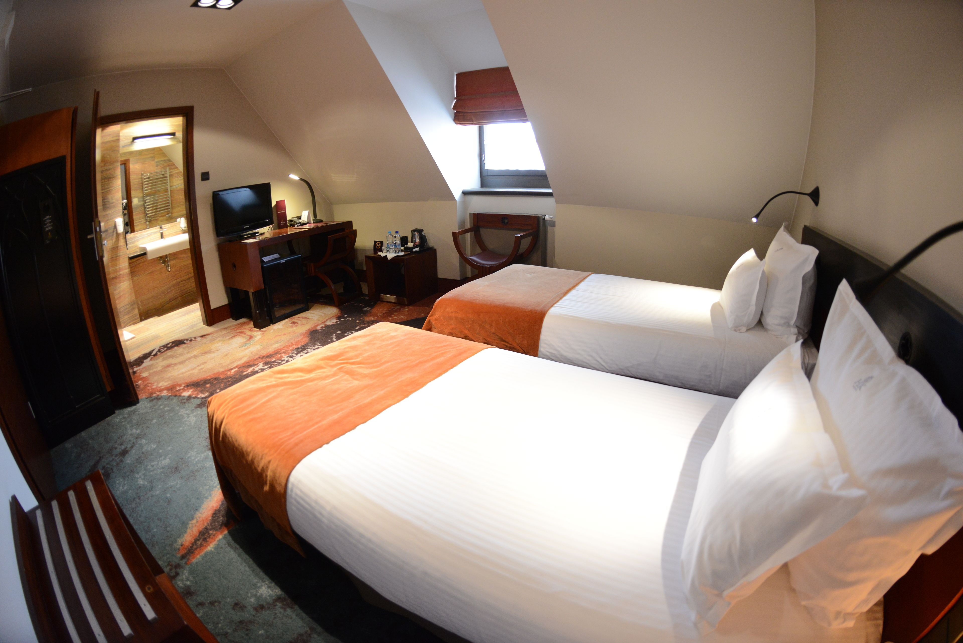 A hotel room with two beds, a desk, a chair, a TV, and a bathroom visible through an open door.