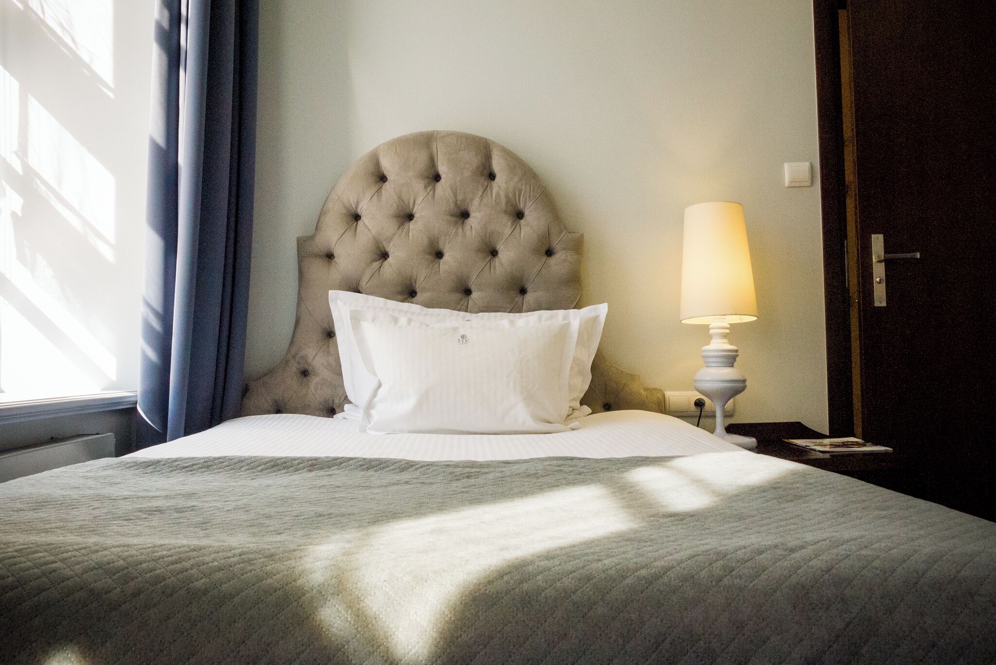 A neatly made bed with a tufted headboard, a bedside lamp, and a window with curtains.