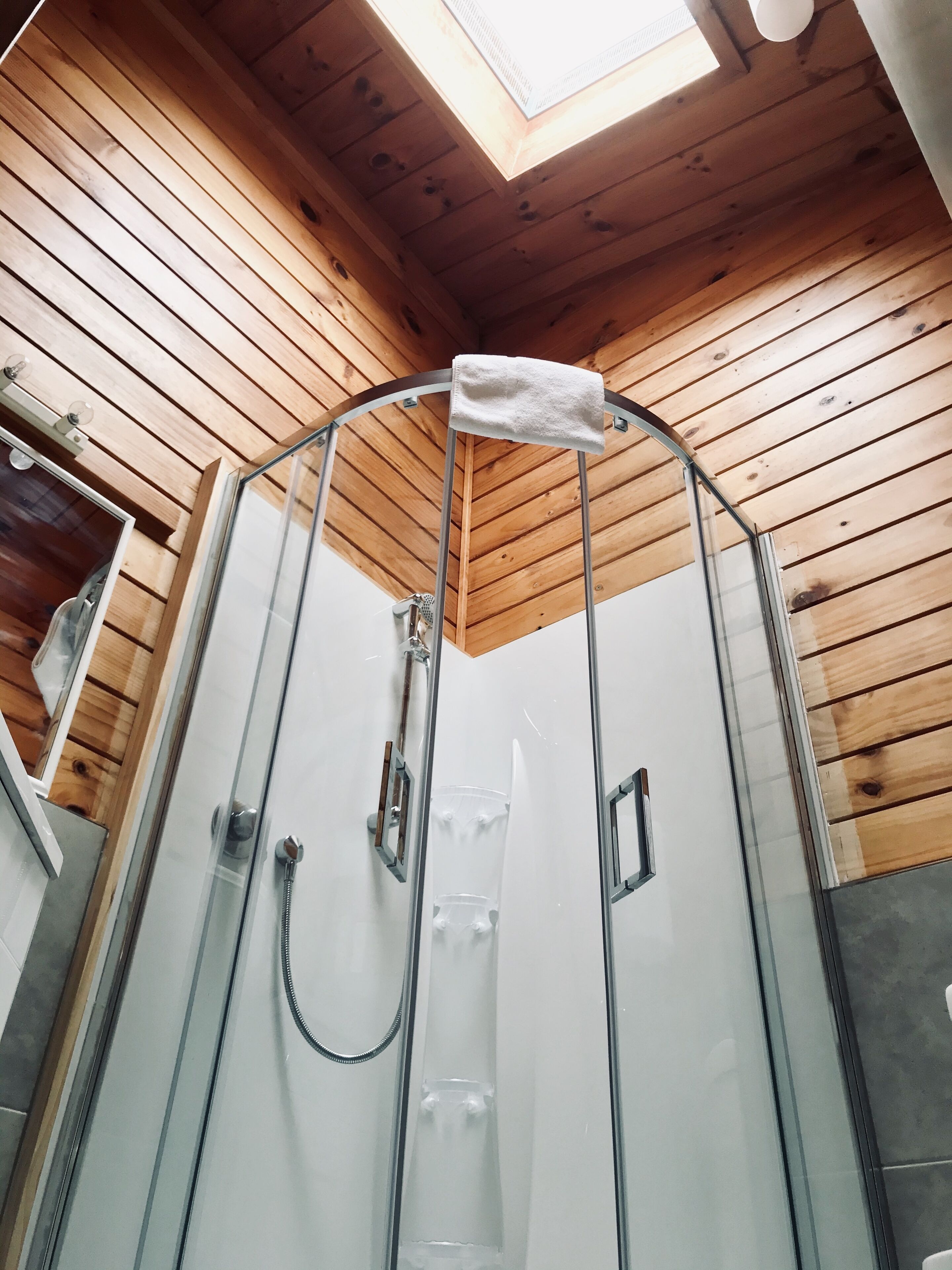 A glass-enclosed shower with a towel hanging on the door, wooden ceiling, and a skylight.