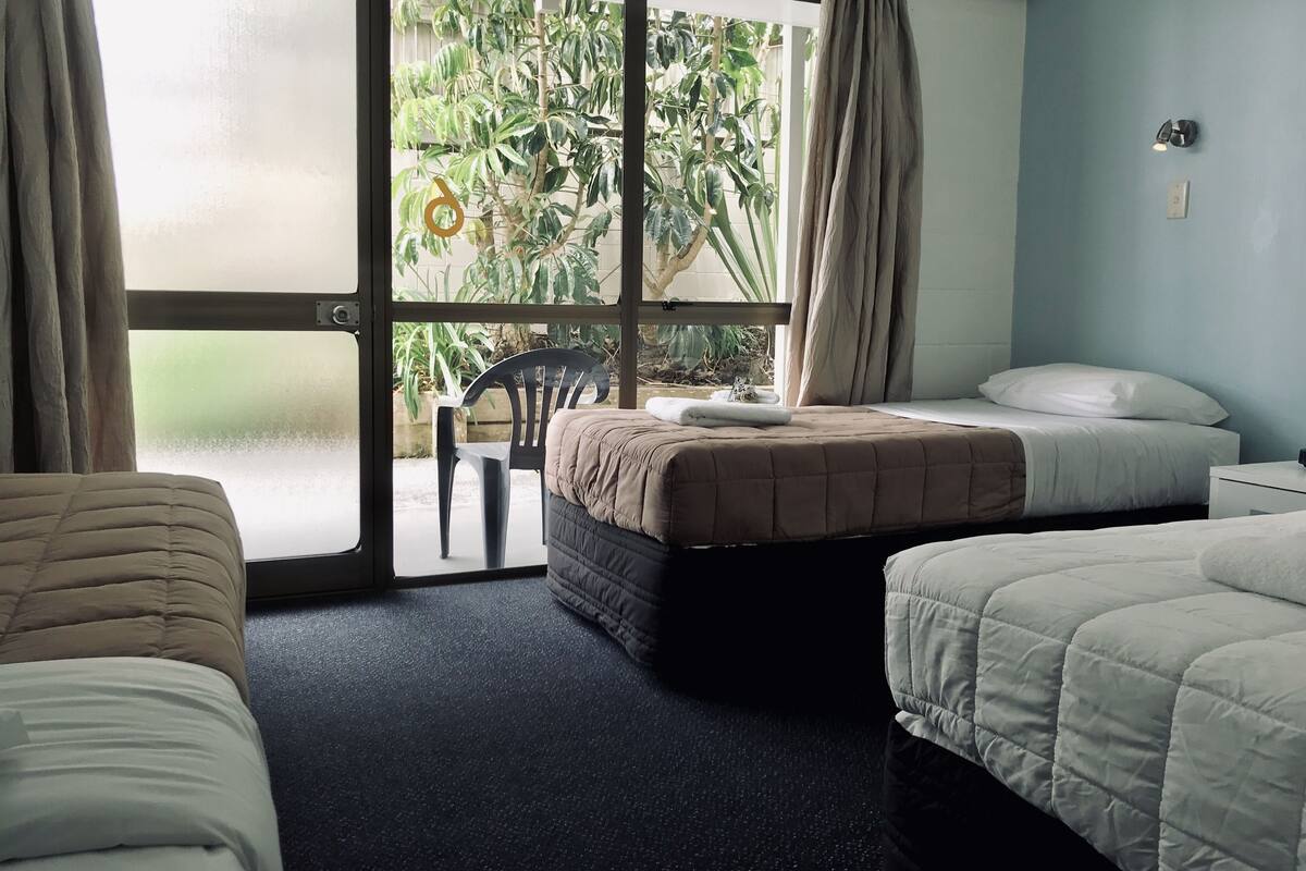 Ein Hotelzimmer mit zwei Betten, einem Sessel und Blick auf Grün hinter einer Glastür.