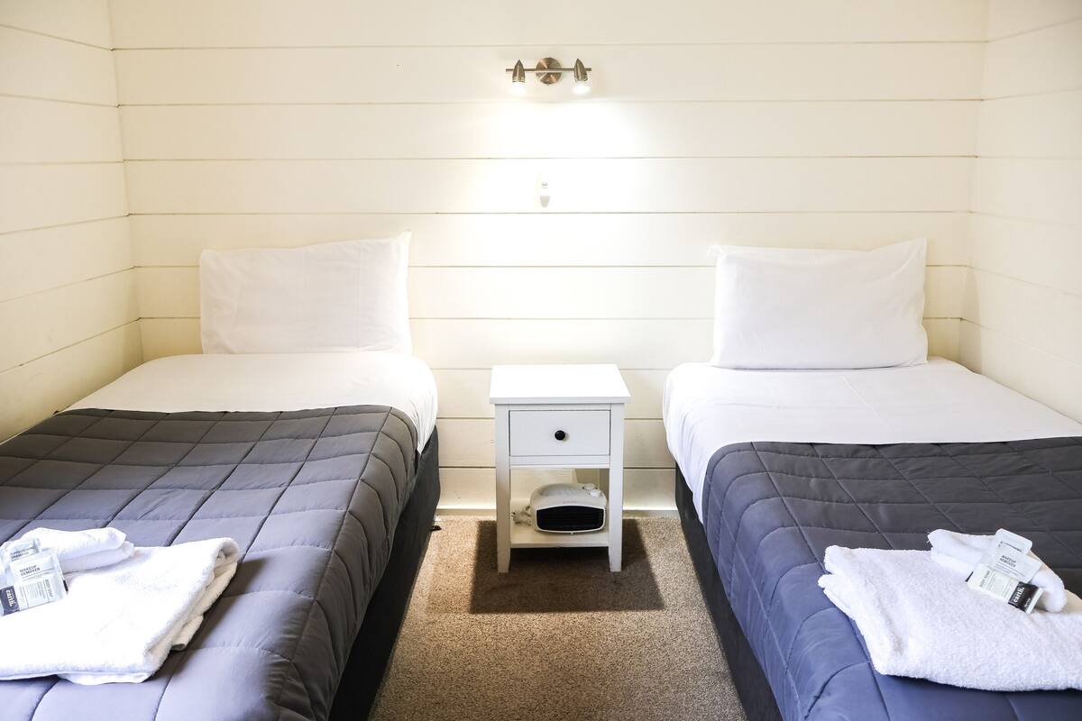 Ein Hotelzimmer mit zwei Betten, jedes mit einem weißen Kopfkissen und einer dunkel gefärbten Bettdecke. Zwischen den Betten befindet sich ein weißer Nachttisch mit einer Schublade und einer Ablagefläche.